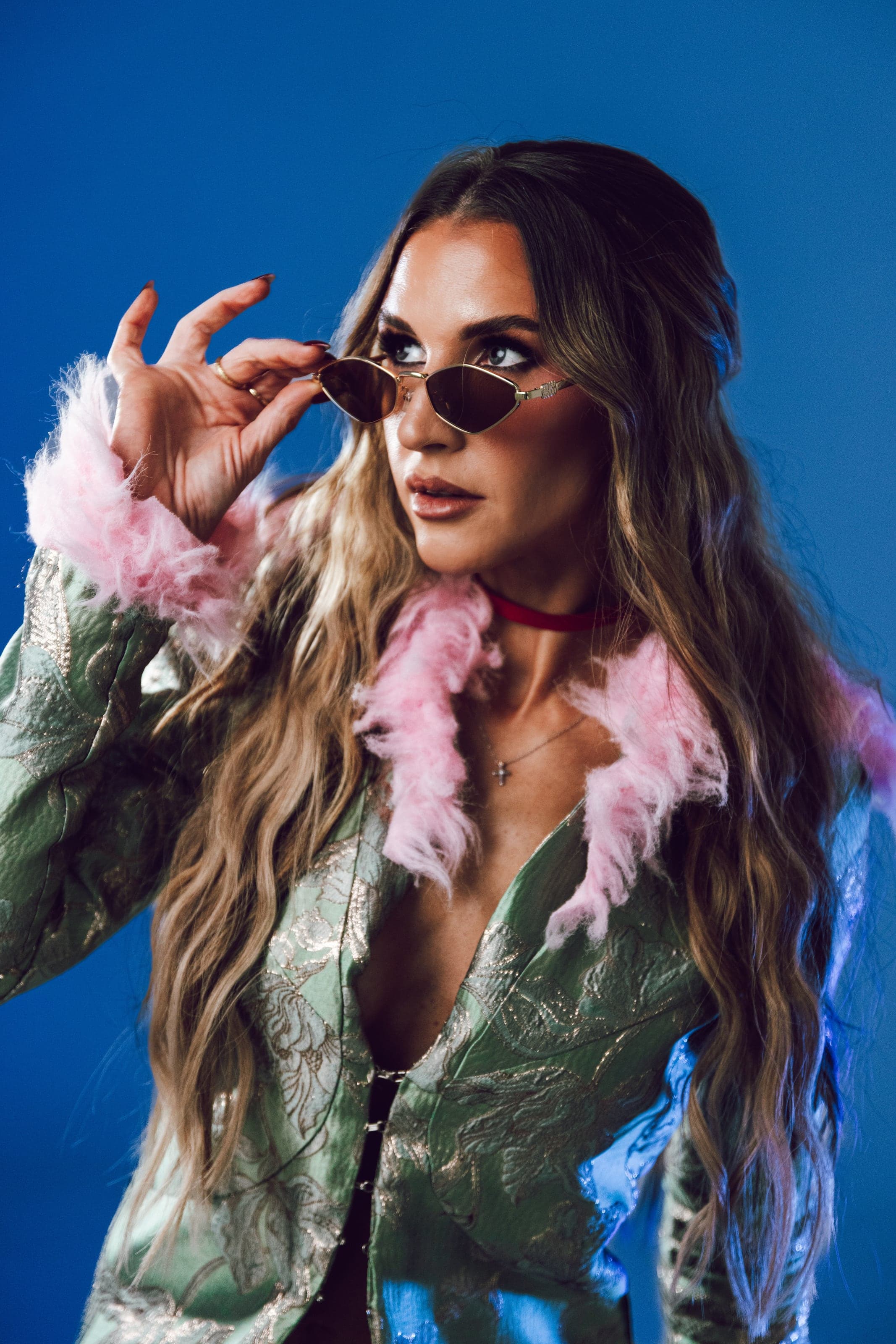 Woman in a brocade coat with pink fur, lowering sunglasses on royal blue